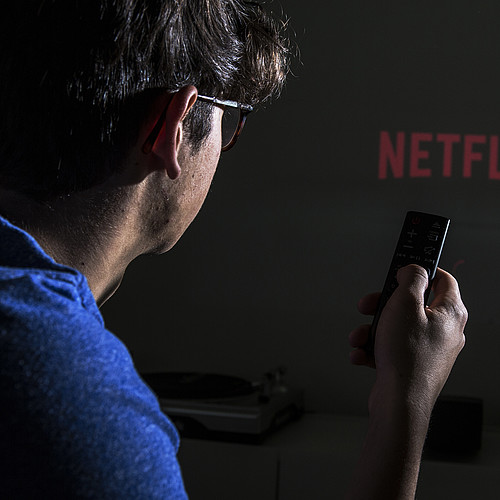 Una persona con un telecomando in mano. Sullo sfondo si può vedere il logo di Netflix.