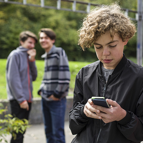 Ein Jugendlicher ist draussen am Handy. Im Hintergrund stehen zwei weitere Jugendliche, die ihn auslachen.