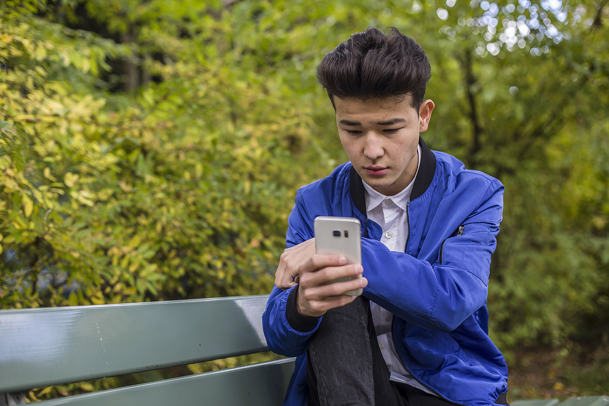 Ein Junge ist draussen und ist am Handy. Er trägt eine blaue Jacke.