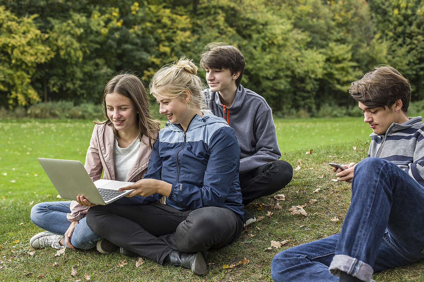 Vier Jugendliche die auf einer Wiese sitzen. Drei davon schauen gemeinsam auf einen Laptop, eine Person auf ein Smartphone.