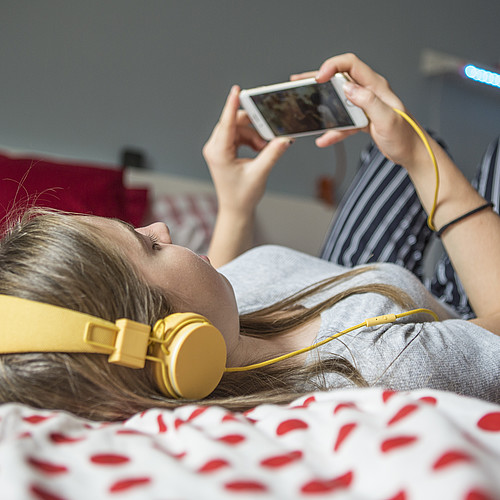 Eine Jugendliche die auf einem Bett liegt und mit Kopfhörer auf dem Smartphone etwas schaut.