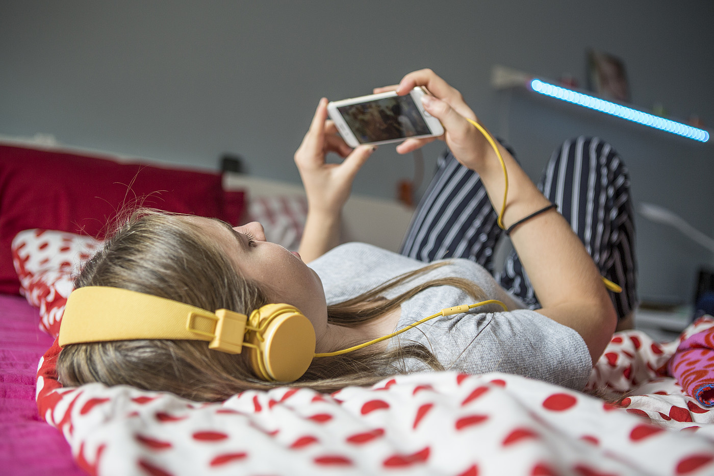 Eine Jugendliche die auf einem Bett liegt und mit Kopfhörer auf dem Smartphone etwas schaut.