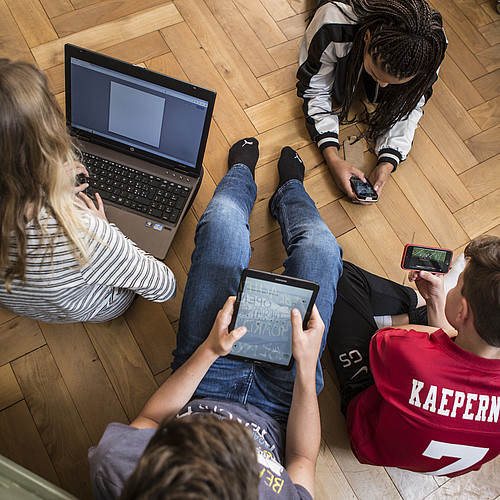 4 Jugendliche die auf dem Boden sitzen oder liegen und alle an einem Laptop, auf einem Tablet oder an einem Smartphone sind.