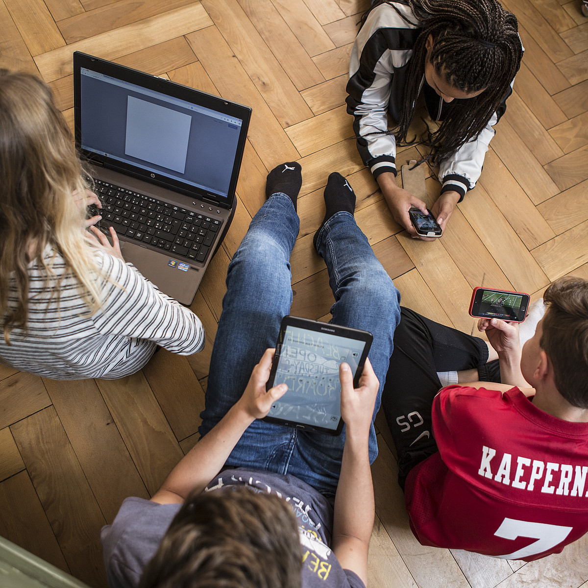 4 Jugendliche die auf dem Boden sitzen oder liegen und alle an einem Laptop, auf einem Tablet oder an einem Smartphone sind.