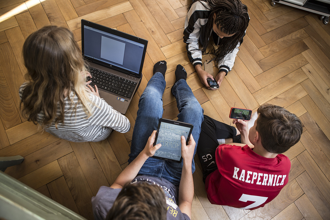 4 Jugendliche die auf dem Boden sitzen oder liegen und alle an einem Laptop, auf einem Tablet oder an einem Smartphone sind.