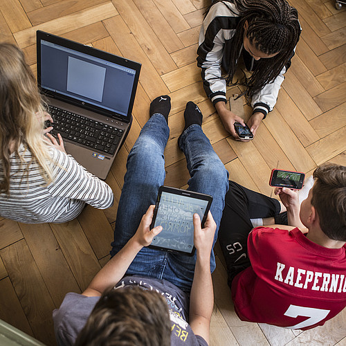 4 Jugendliche die auf dem Boden sitzen oder liegen und alle an einem Laptop, auf einem Tablet oder an einem Smartphone sind.