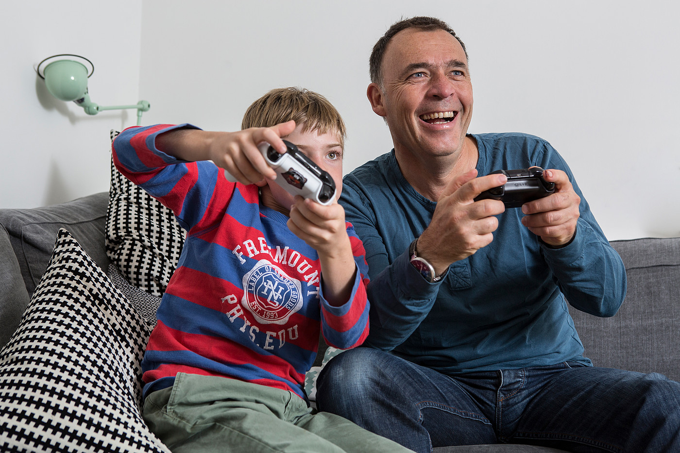 Ein Vater der mit seinem Sohn auf dem Sofa sitzt und am Videospiele spielen ist.