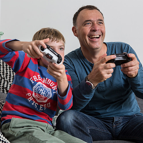 Ein Vater der mit seinem Sohn auf dem Sofa sitzt und am Videospiele spielen ist.