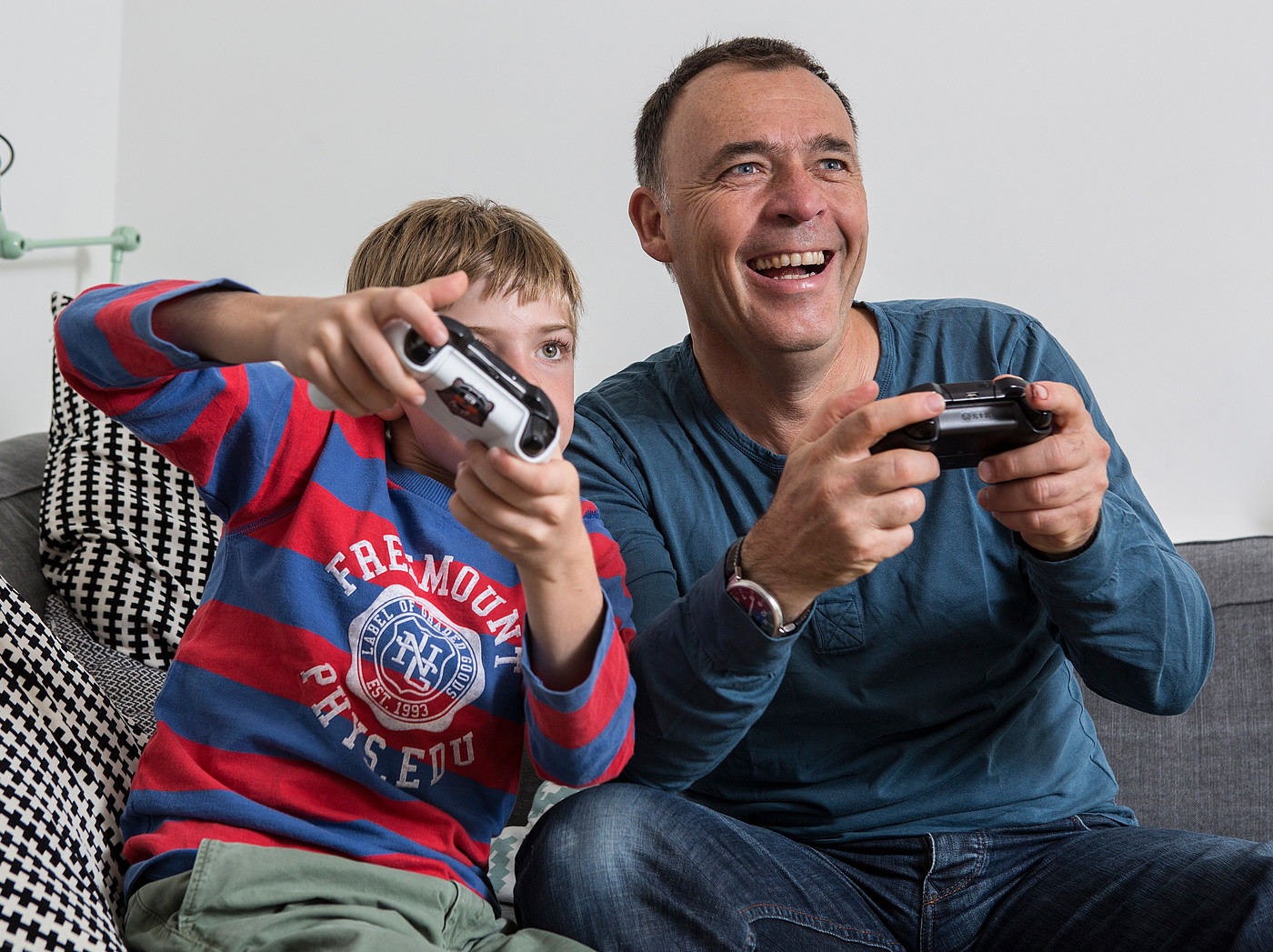 Ein Vater der mit seinem Sohn auf dem Sofa sitzt und am Videospiele spielen ist.