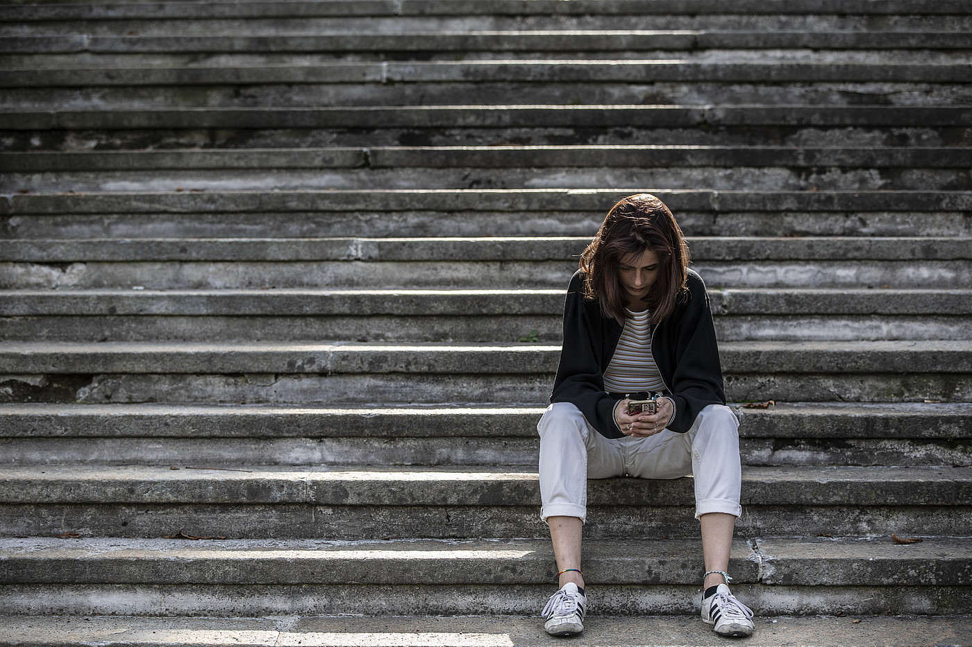 Junge Frau sitzt alleine auf einer Treppe und blickt in ihr Smartphone
