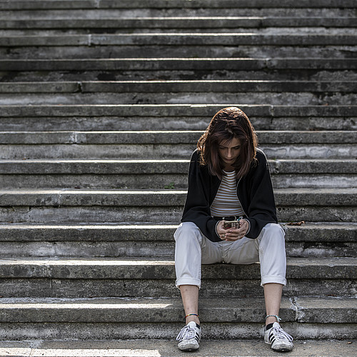 Junge Frau sitzt alleine auf einer Treppe und blickt in ihr Smartphone
