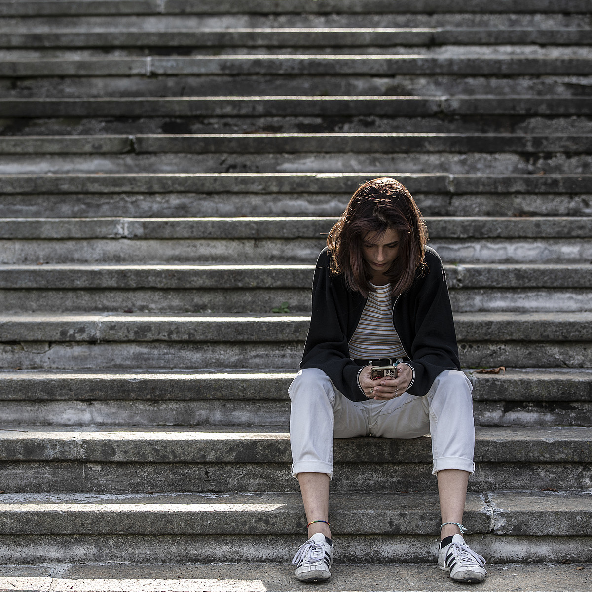 Junge Frau sitzt alleine auf einer Treppe und blickt in ihr Smartphone