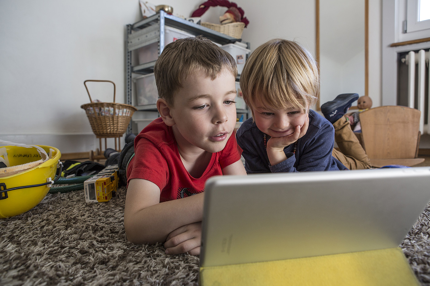 Zwei Kinder die in einem Kinderzimmer auf dem Boden liegen und auf ein Tablet schauen.