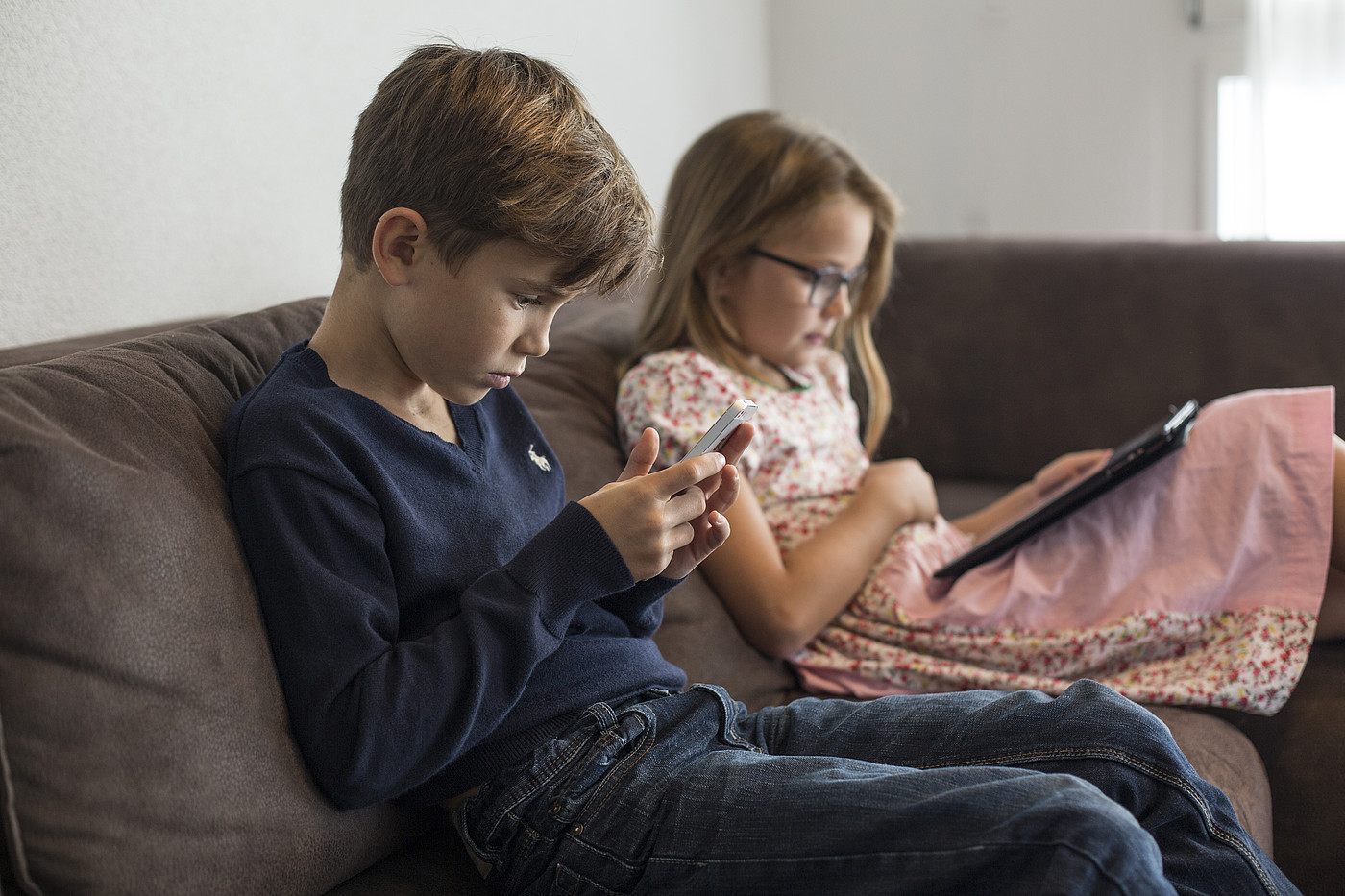 Zwei Kinder die mit einem Smartphone oder einem Tablet in der Hand auf einem Sofa sitzen.