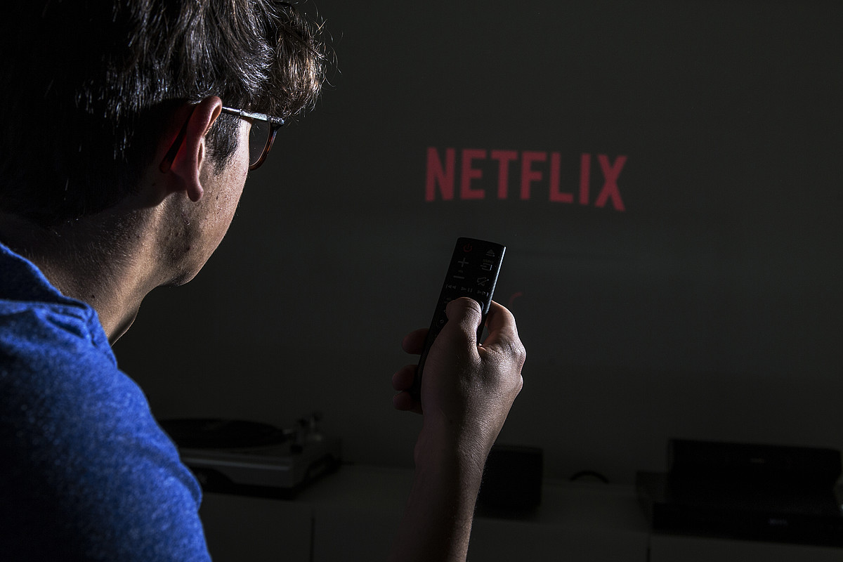 Eine Person mit einer Fernbedienung in der Hand. Im Hintergrund ist das Netflix logo zu erkennen.