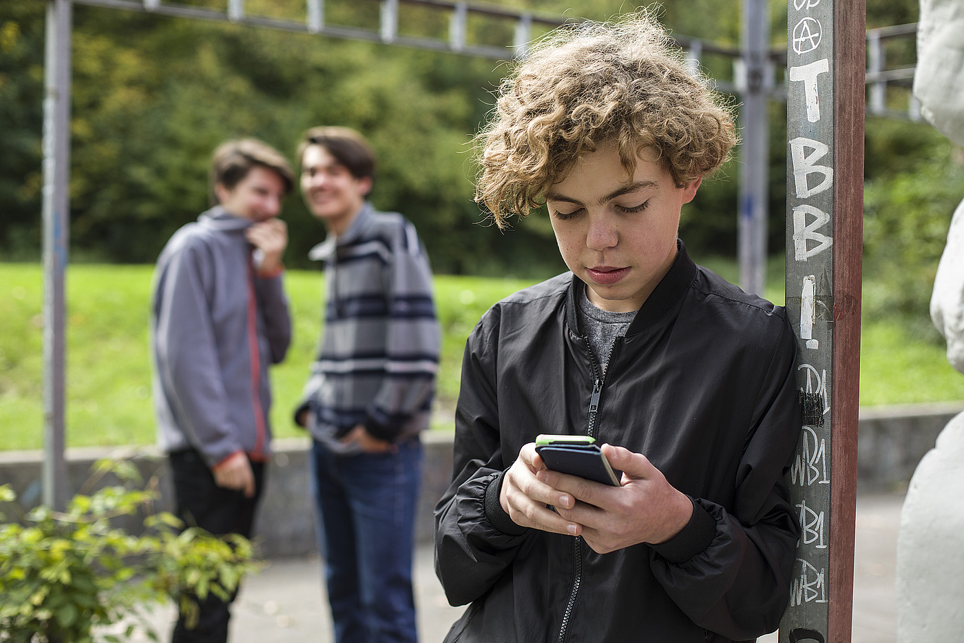 Ein Jugendlicher ist draussen am Handy. Im Hintergrund stehen zwei weitere Jugendliche, die ihn auslachen.