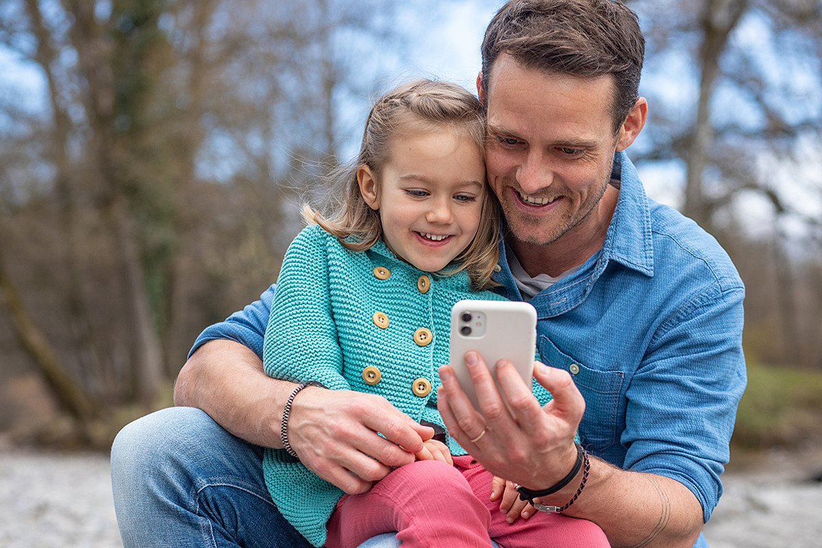 Ein Vater mit einem Kind auf den Knien die gemeinsam auf ein Smartphone schauen.