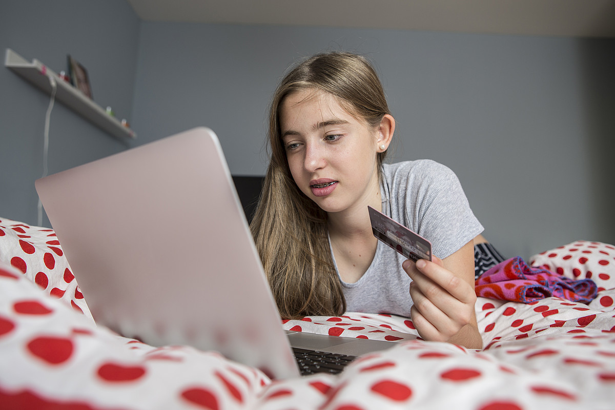 Eine Jugendliche liegt mit dem Laptop auf dem Bett und kauft sich etwas mit der Kreditkarte.