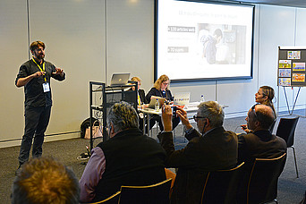 Ein Vortragender vor Publikum. Ein Zuhörer macht ein Foto von der Präsentation.