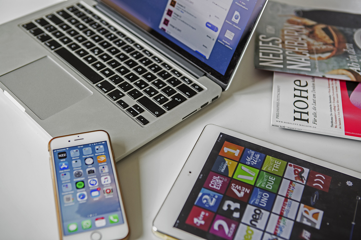 Ein Laptop, ein Smartphone, ein Tablet und Zeitschriften die auf einem Tisch liegen.