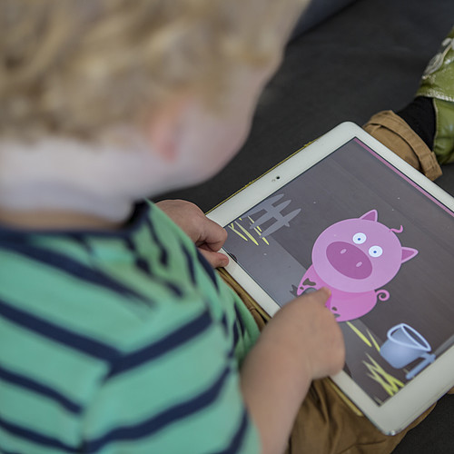 Ein kleiner Junge schaut sich etwas auf dem Tablet an. Auf dem Tablet ist ein animiertes Schwein zu sehen.