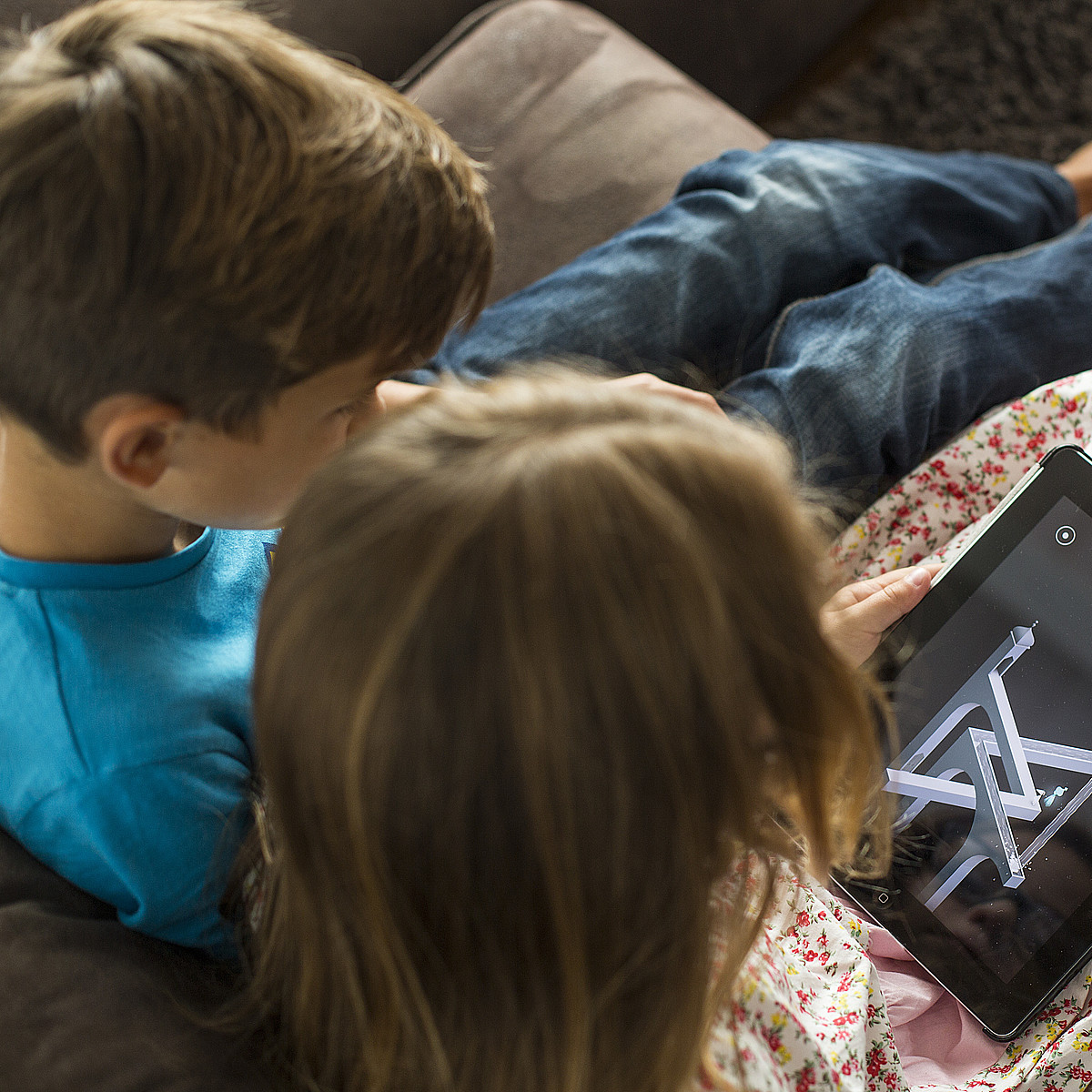 Zwei Kinder die auf einem Sofa sitzen und auf ein Tablet schauen.