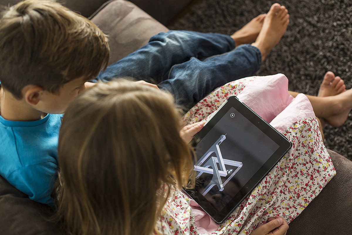 Zwei Kinder die auf einem Sofa sitzen und auf ein Tablet schauen.