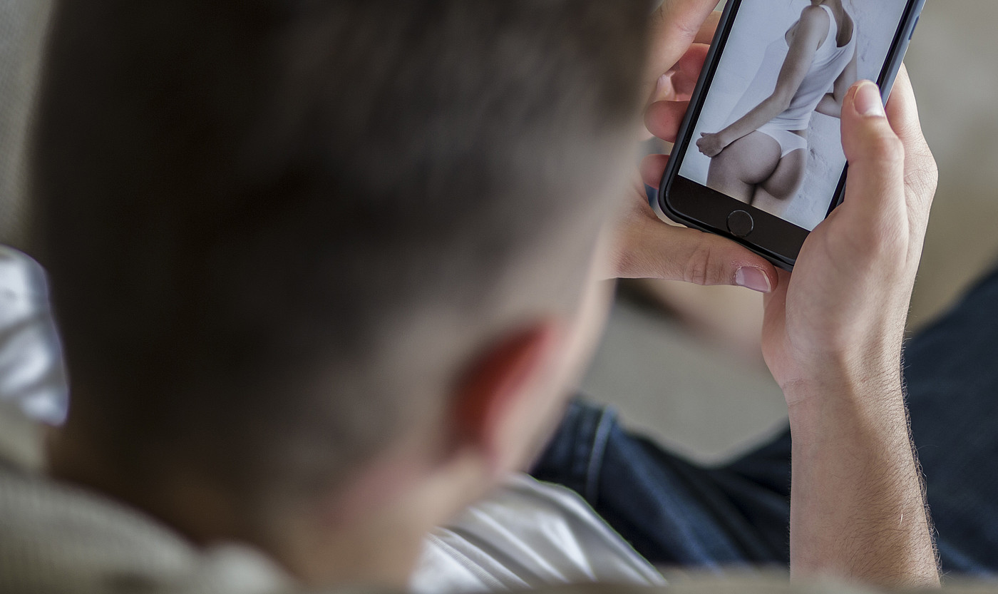 Ein junger Mann schaut sich ein Bild einer halbnackten Frau auf dem Handy an. 